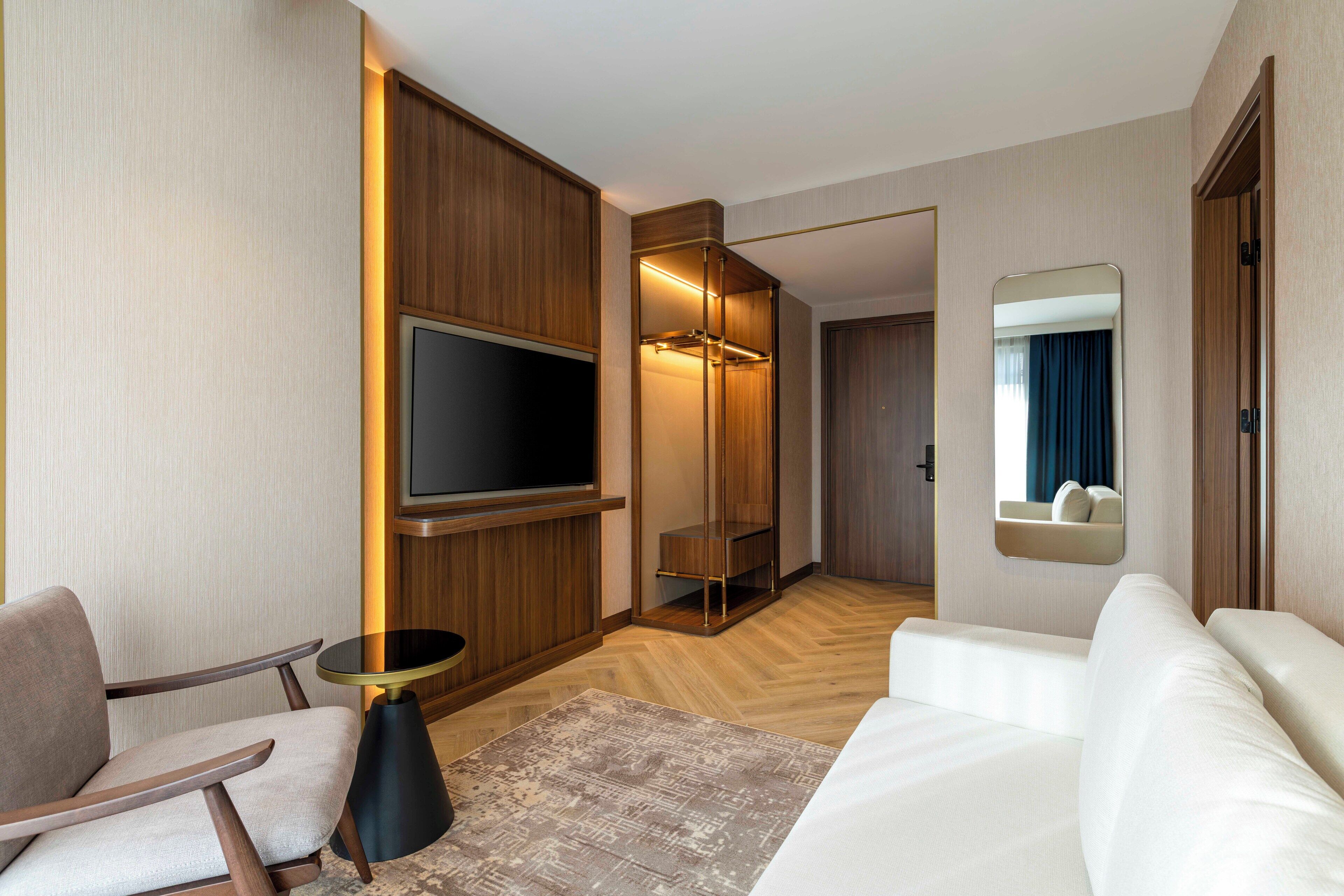 A modern hotel room with a flat-screen TV mounted on the wall, a seating area with a sofa and armchairs, a small round table, and a mirror above the mirror cabinet.