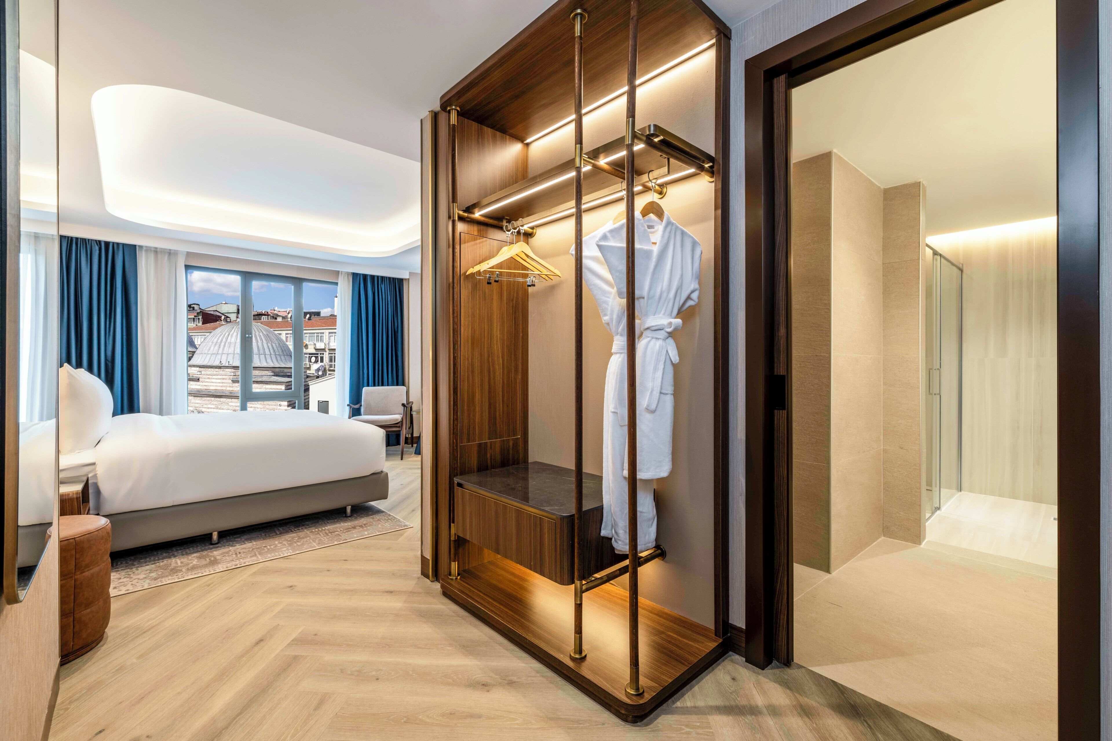 A modern hotel room with a large bed, a walk-in closet with a robe hanging, and a city view through the window.