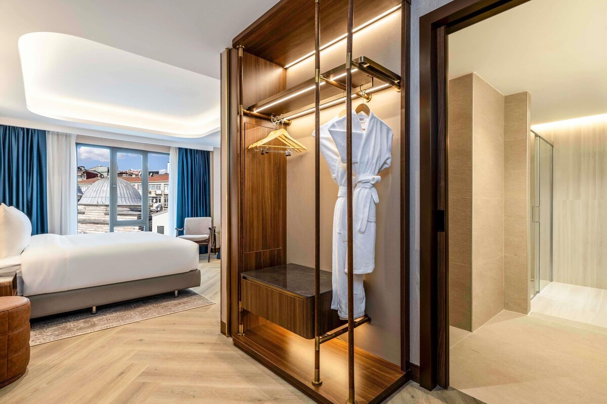 A modern hotel room with a large bed, a walk-in closet with a robe hanging, and a view of buildings through the window.