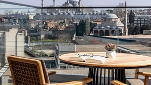 A balcony with a view of a mosque with multiple domes and minarets, a table with a book and glasses, and wicker chairs. - Mövenpick Istanbul Old Town (Istanbul)