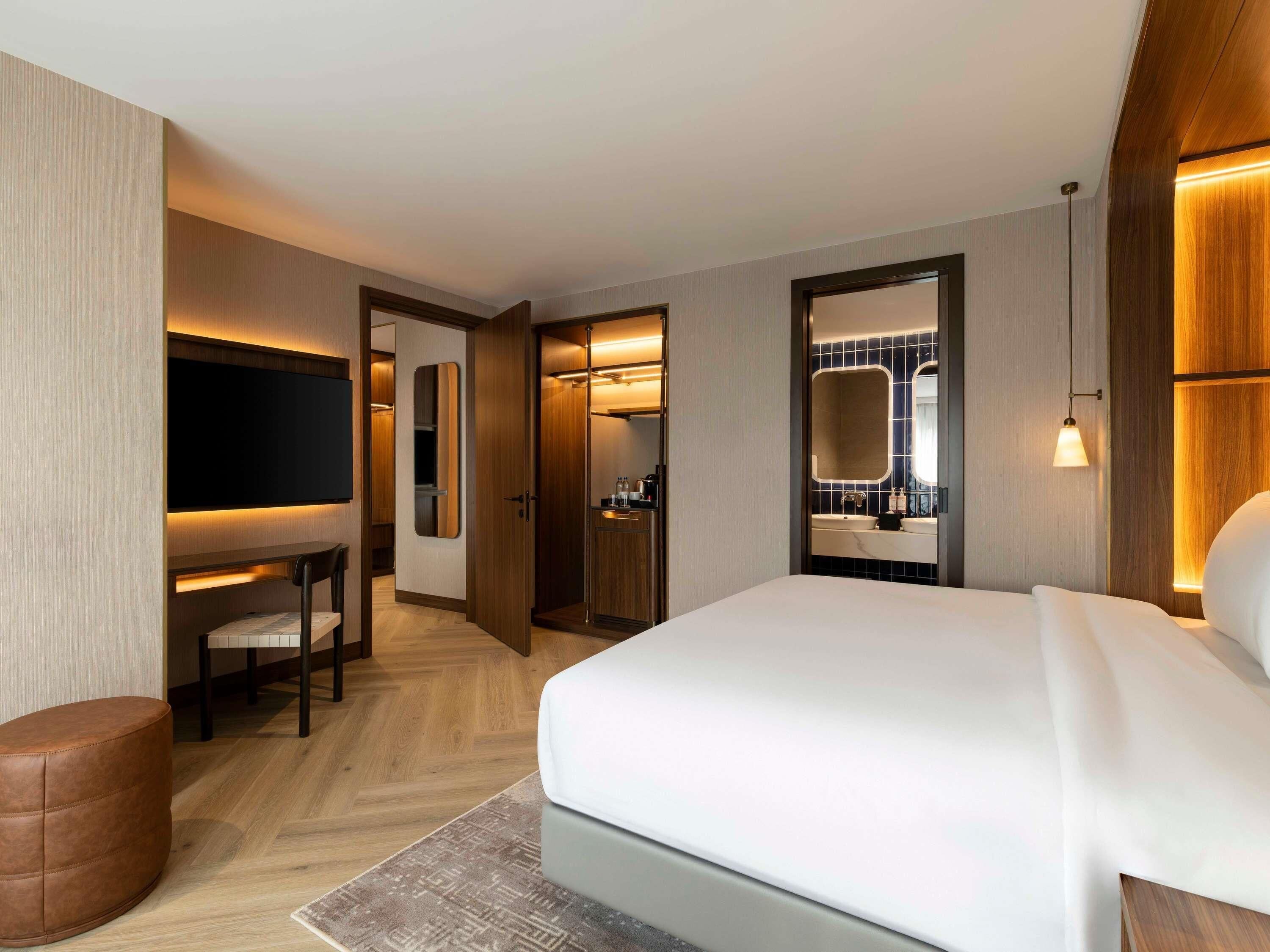 A hotel room with a bed, a desk with a chair, a TV, a bathroom with a mirror, and a small seating area.
