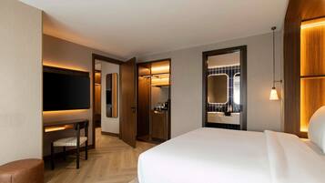 A hotel room with a bed, a desk with a chair, a TV, a bathroom with a mirror, and a small seating area.