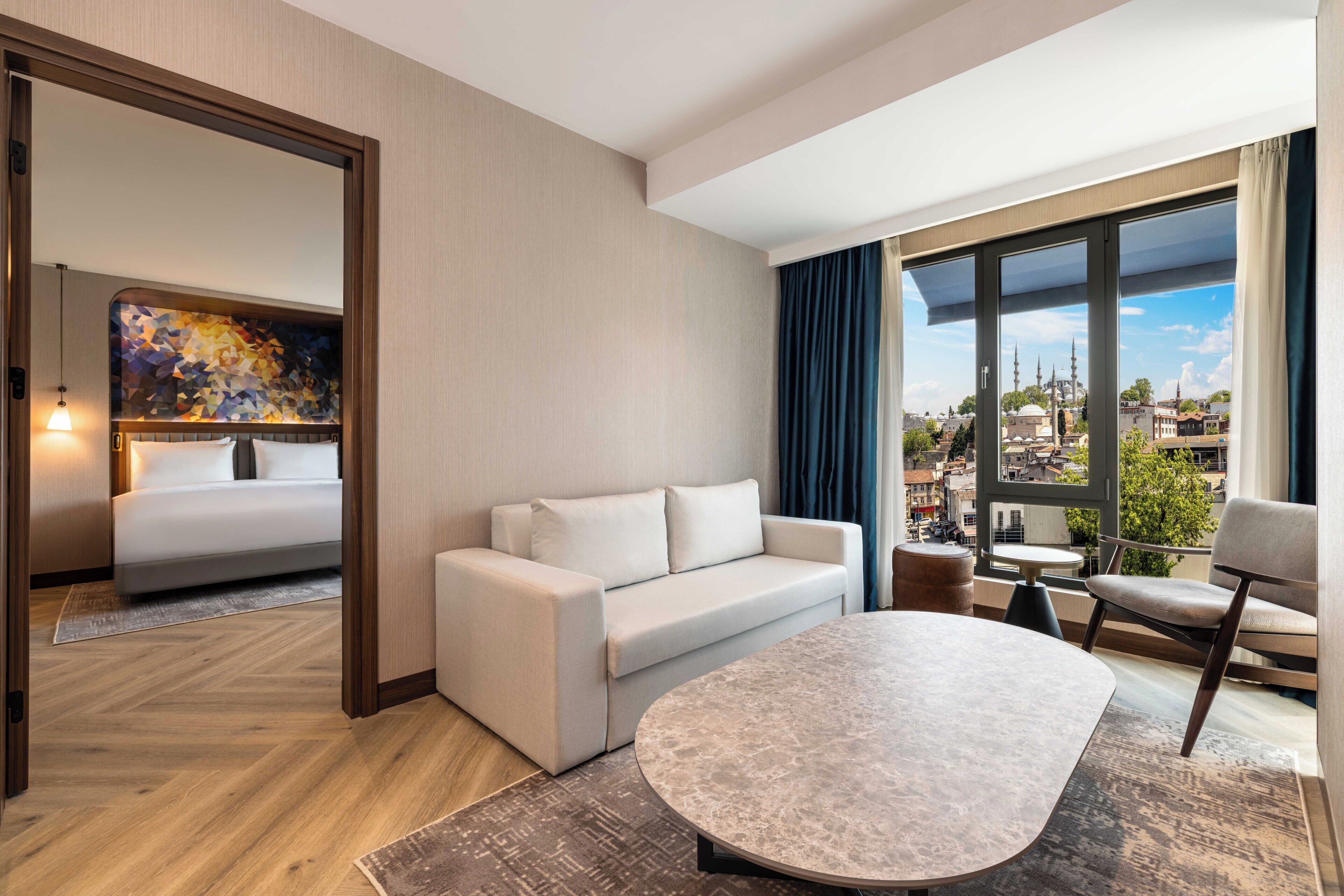 A modern hotel room with a sofa, a round table, a view of the city, and a large window.