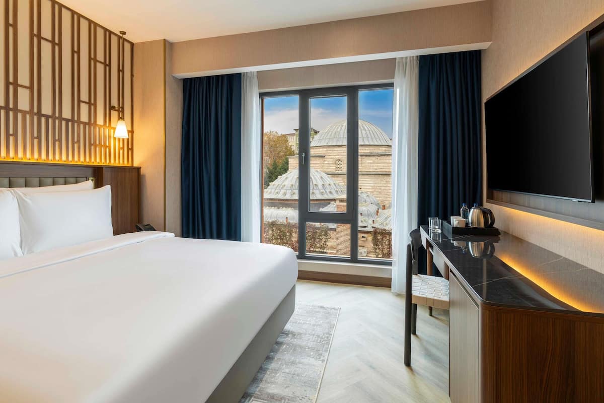 Habitación de hotel con una cama grande, un escritorio con televisor de pantalla plana, vista a un edificio con cúpulas y un ventanal con cortinas.