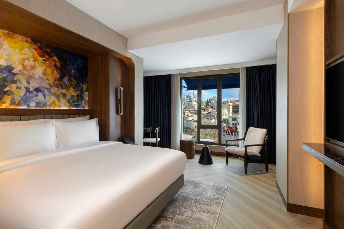 Habitación de hotel con una cama grande, un escritorio, una silla, un televisor y un ventanal con vistas a la ciudad.