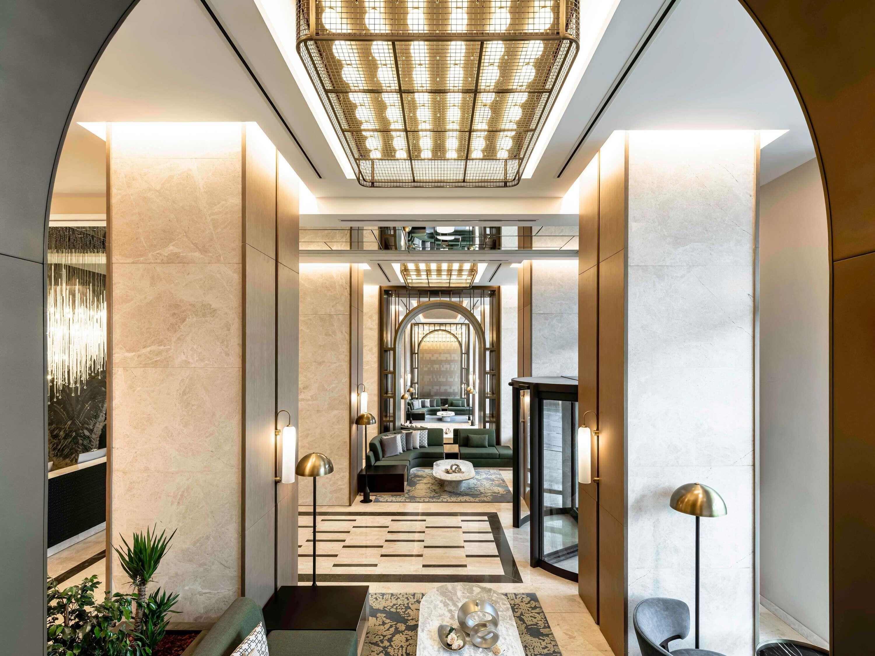 A modern hotel lobby with a central seating area, a chandelier, and a view into the dining area.
