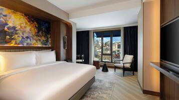 A hotel room with a large bed, a flat-screen TV, a painting on the wall, and a view of the city through the window.