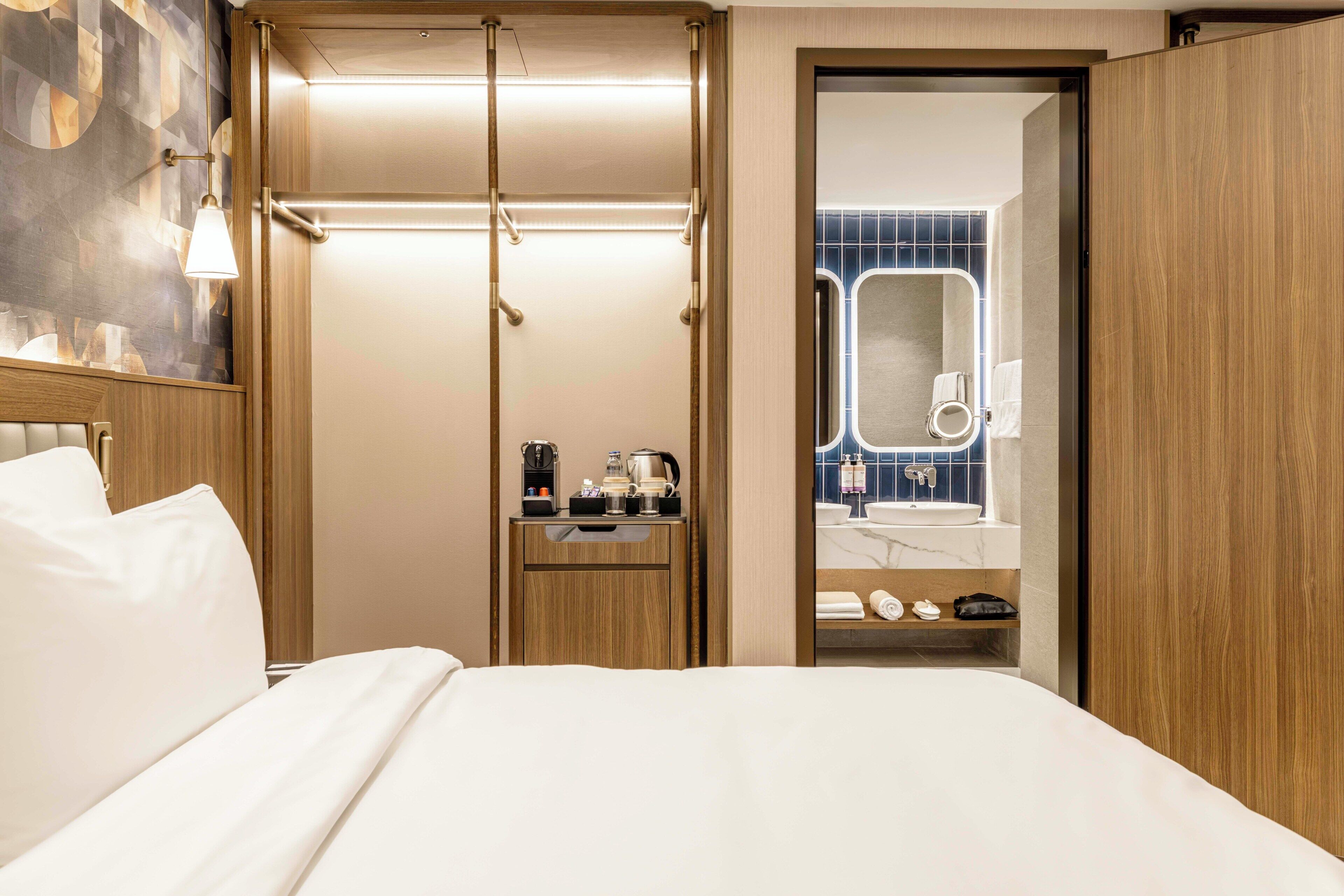 A hotel room with a bed, a wooden wardrobe, a small bedside table with a coffee maker, and a bathroom visible through an open door.