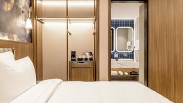 A hotel room with a bed, a wooden wardrobe, a small bedside table with a coffee maker, and a bathroom visible through an open door.