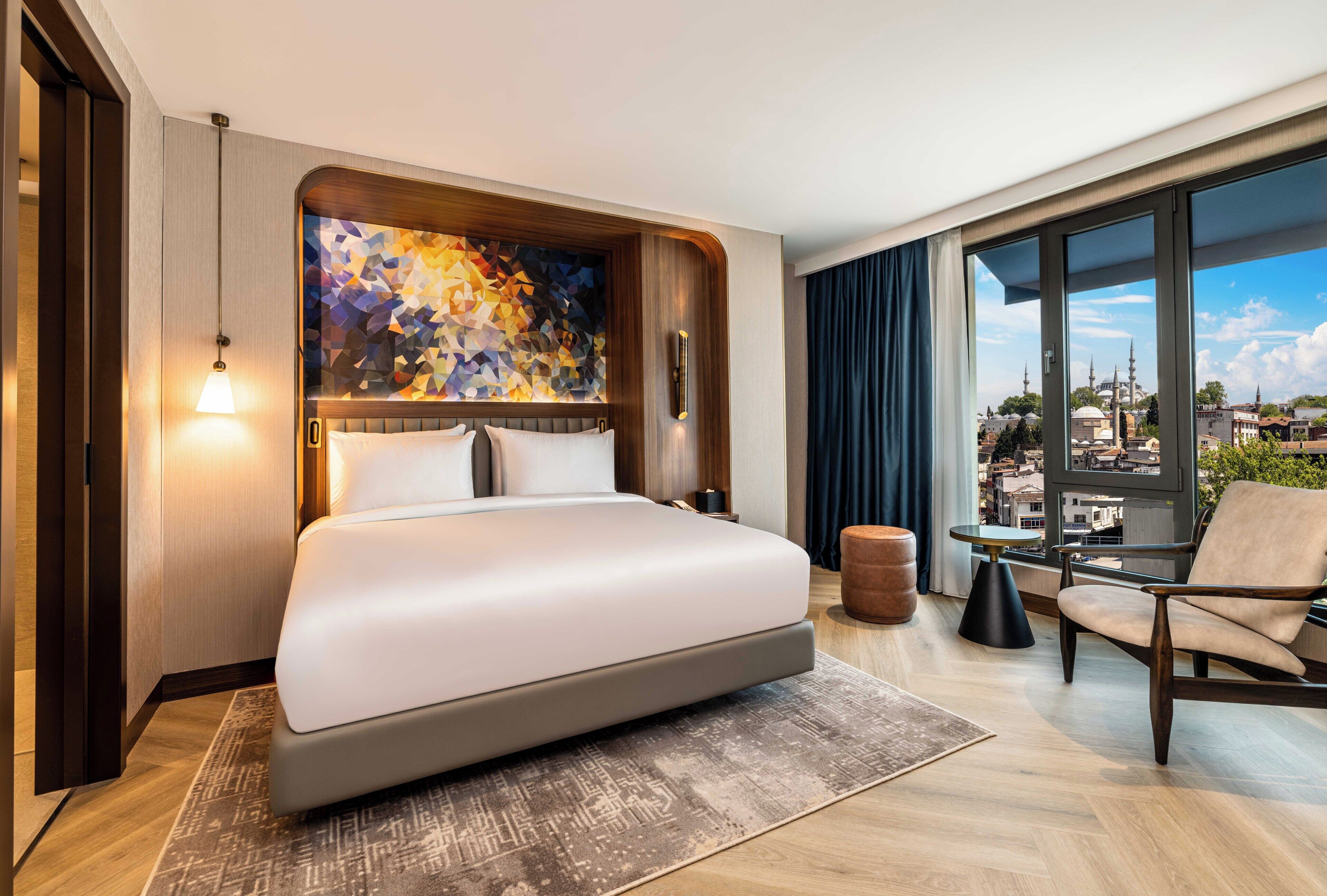 Ein modernes Hotelzimmer mit einem großen Bett, Blick auf die Stadt und einem kleinen Tisch mit Stuhl.