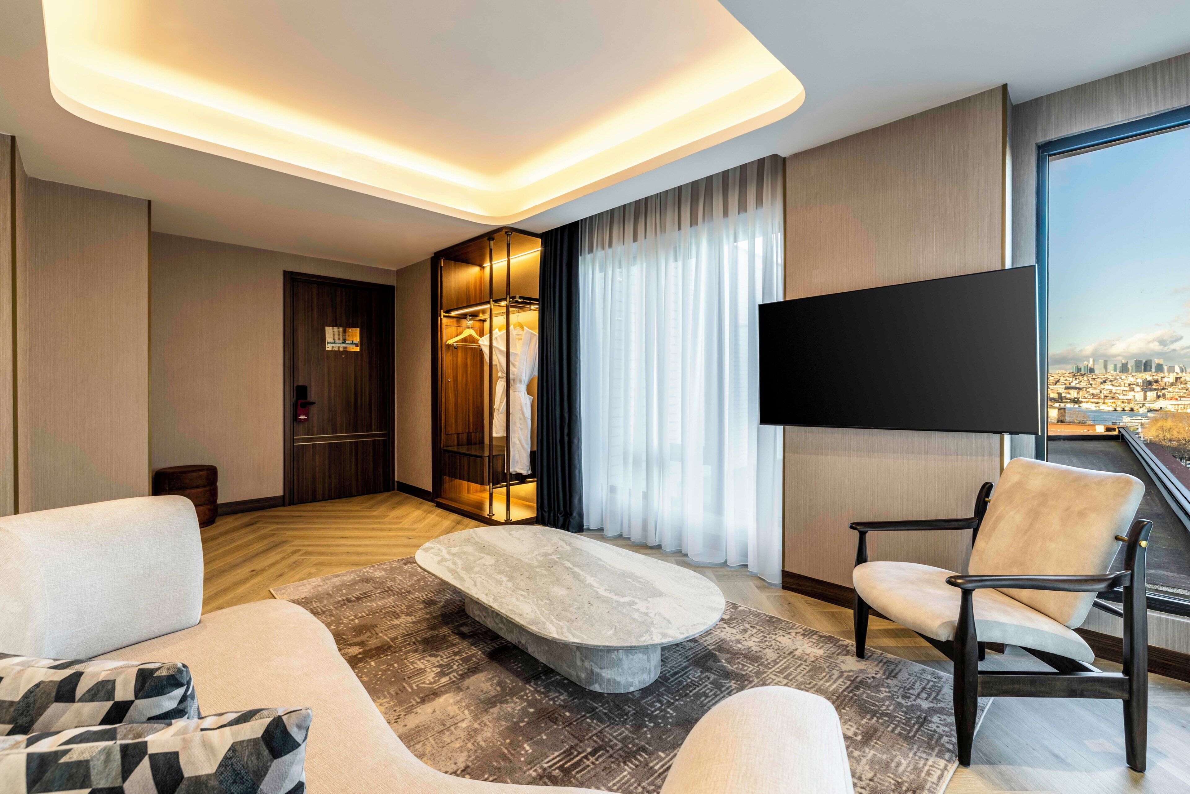 A modern hotel room with a large flat-screen TV, a stone coffee table, a sofa, a chair, a wardrobe, and a city view.