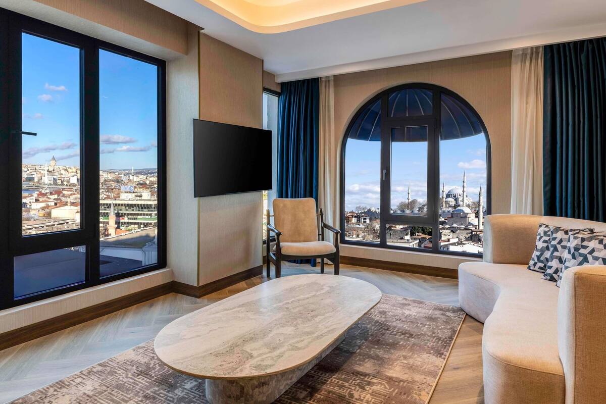 Habitación de hotel moderna con un ventanal grande que ofrece vistas a la ciudad, un televisor de pantalla plana montado en la pared, una mesa de centro redonda y un sofá con cojines estampados.