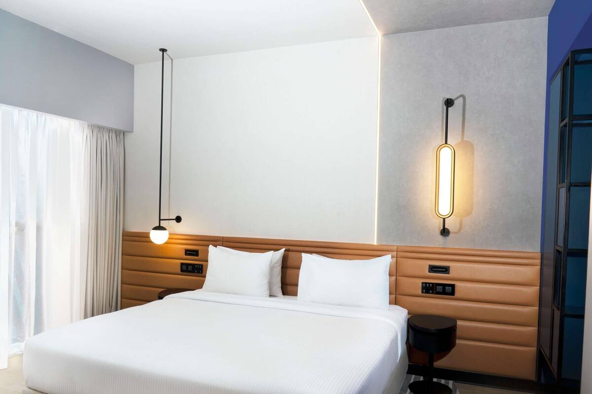 Habitación de hotel con una cama grande, una mesita de noche, una lámpara de pared y una ventana con cortinas.