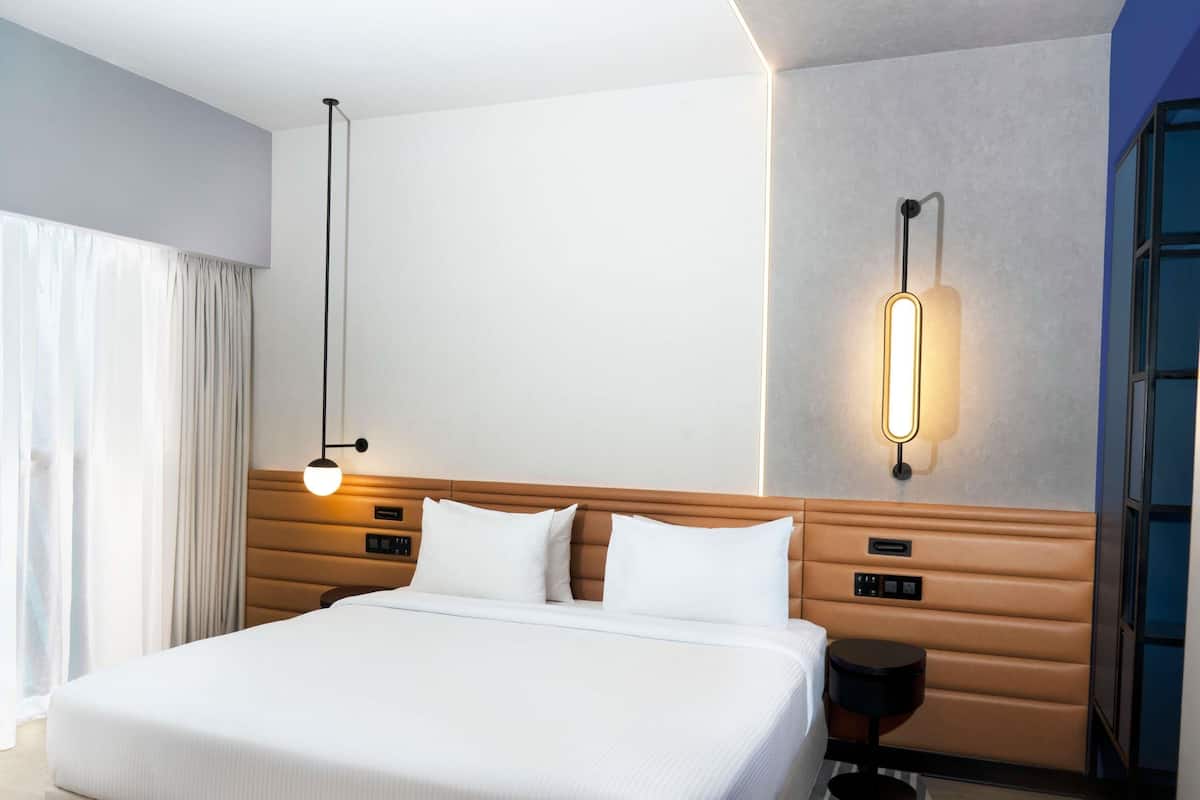 A hotel room with a large bed, a bedside table, a wall-mounted light, and a window with curtains.