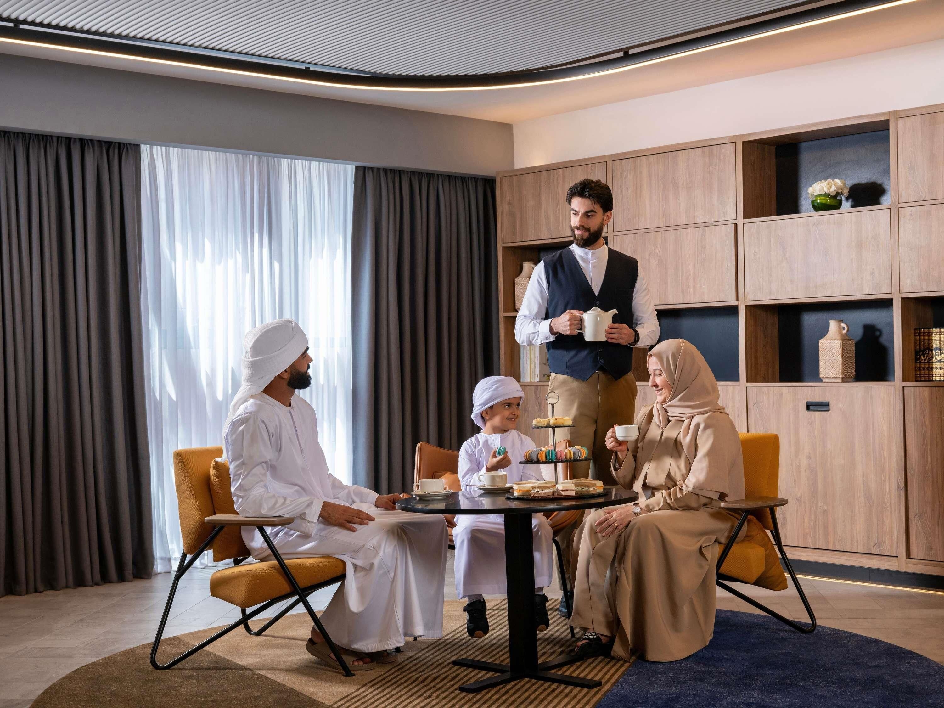 A modern hotel room with a round table, wooden shelving, and a large window with curtains.