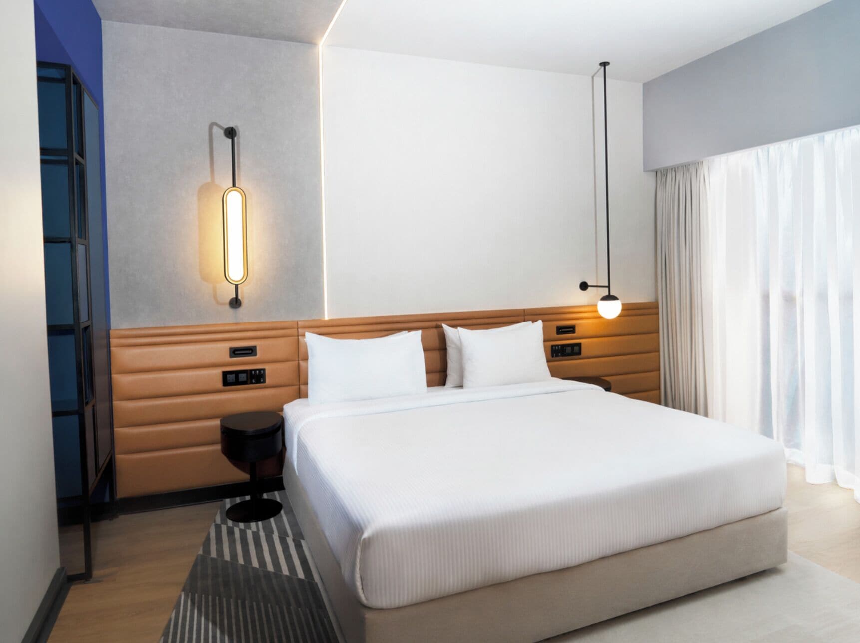 Quarto de hotel moderno com uma cama grande, mesas de cabeceira, um criado-mudo, uma luminária fixada na parede e uma janela com cortinas.