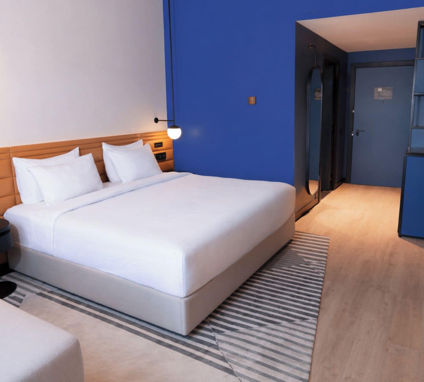 Une chambre d’hôtel avec un grand lit, une table de chevet, une commode, une lampe, une porte et un petit bureau.