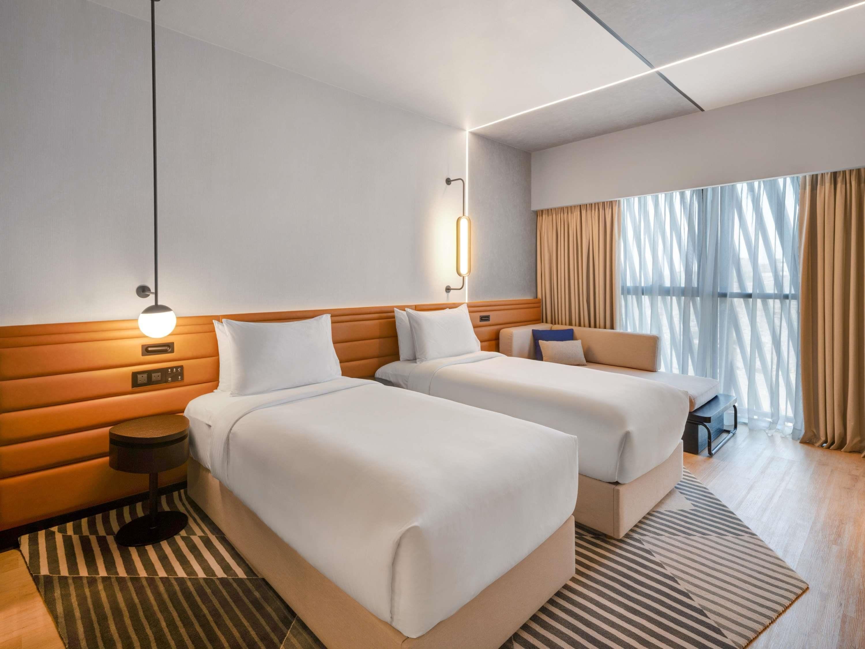 Ein Hotelzimmer mit zwei Betten, einem Nachttisch, einer Lampe und einem großen Fenster mit Vorhängen.