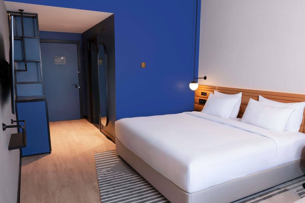 Una habitación de hotel con cama, mesita de noche, lámpara, espejo, armario y puerta.