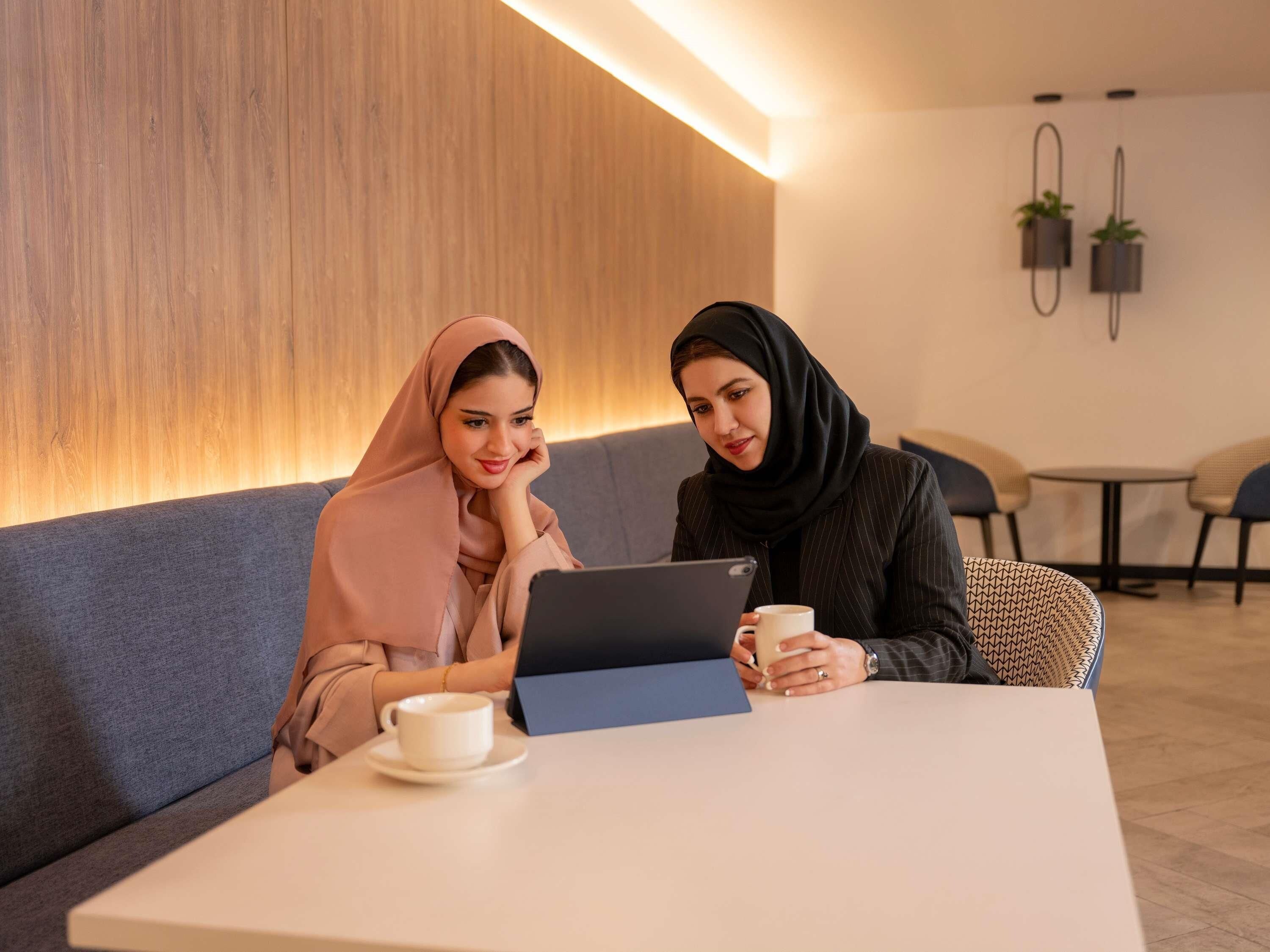 Two individuals seated at a table with a laptop and a cup, in a room with a wooden wall and a small table with chairs in the background.