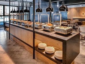 A buffet area with various food items, a counter with plates, and a seating area with tables and chairs. - Novotel Madinah (Madinah)