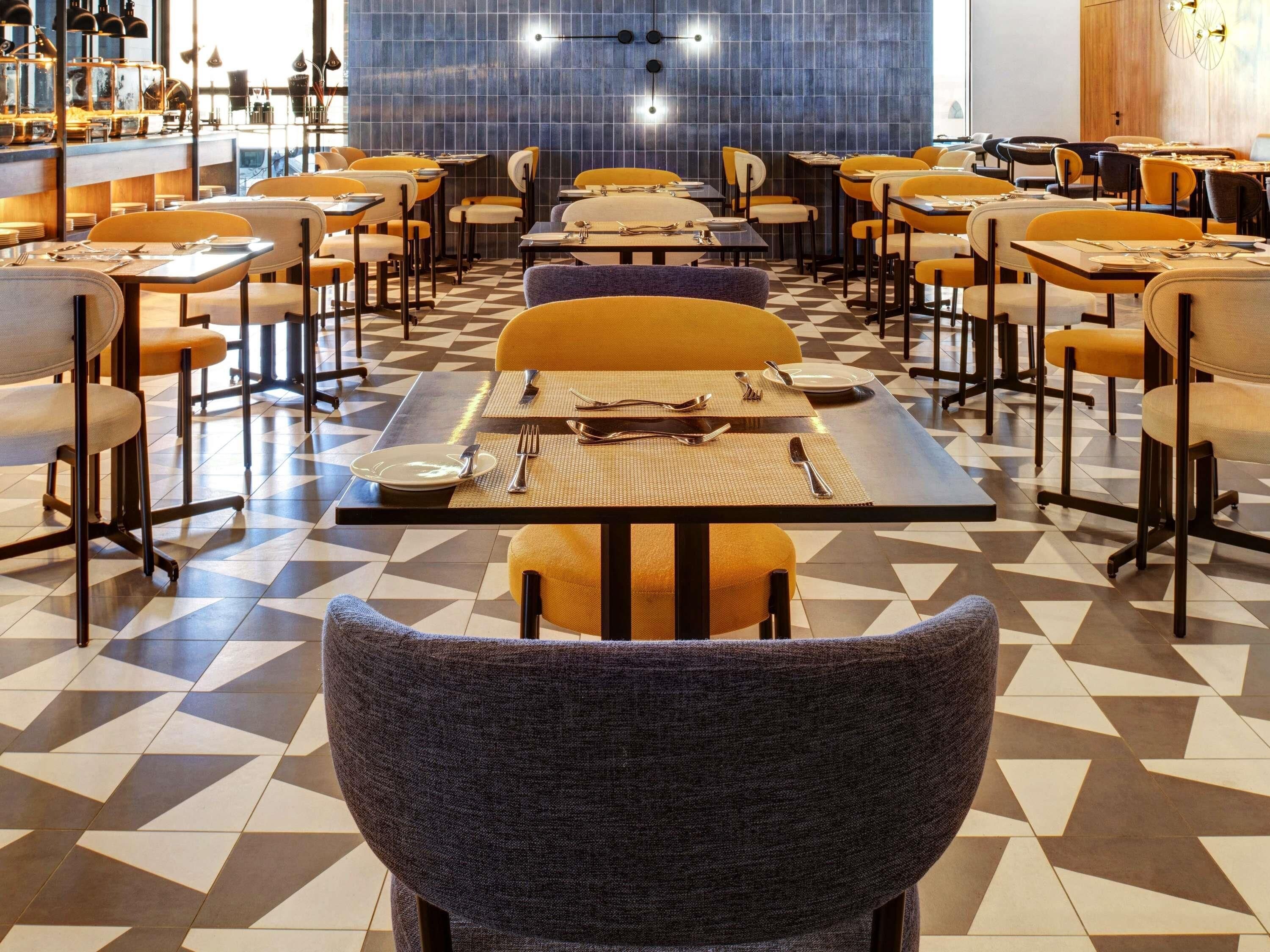 A restaurant with a geometric patterned floor, tables with yellow and grey chairs, and a blue-tiled wall.