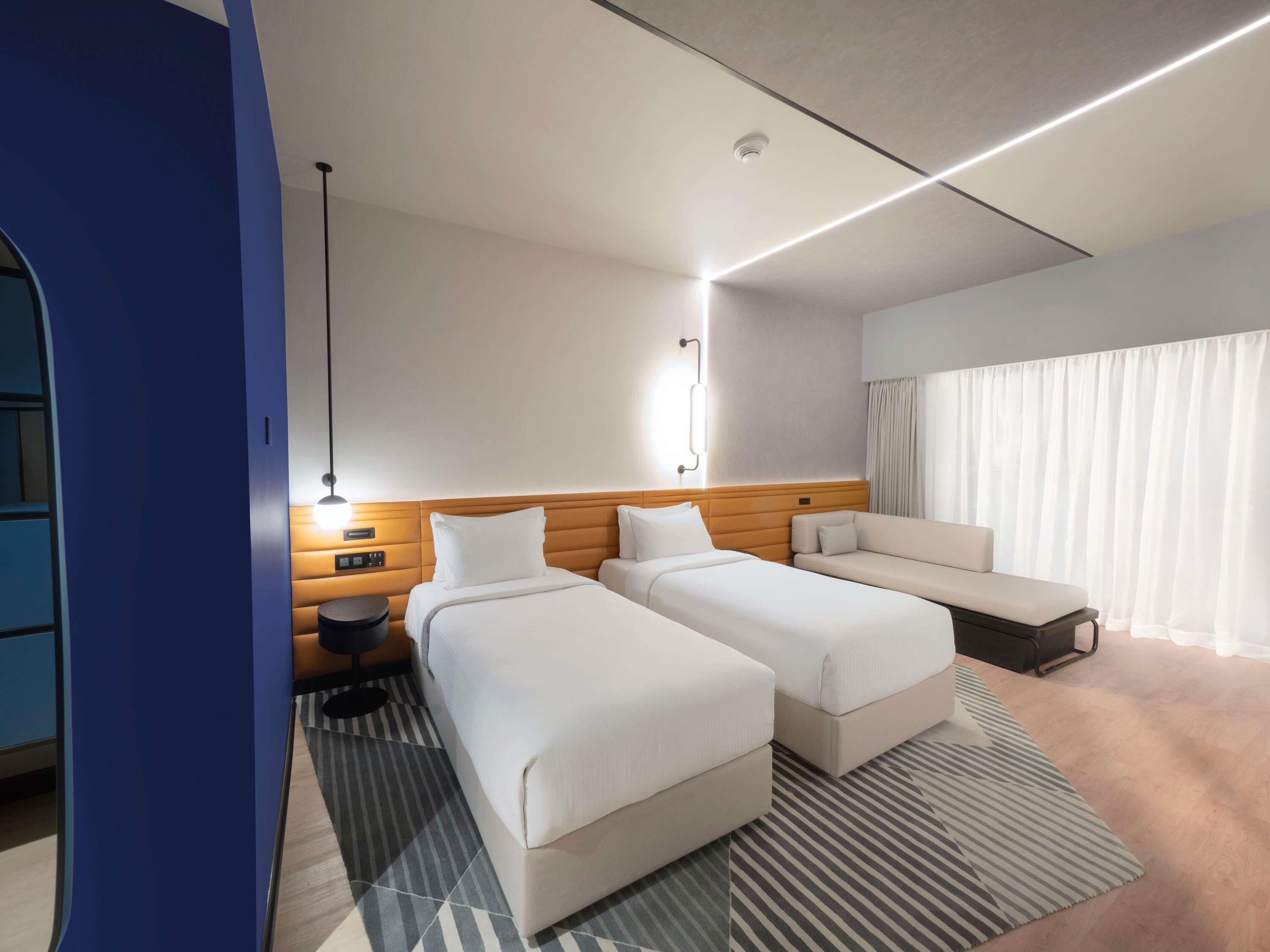 A hotel room with two beds, a sofa, a desk, and a large window with curtains.