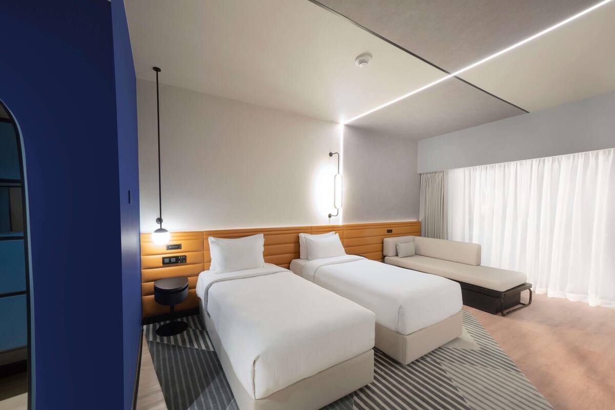 Habitación de hotel con dos camas, un sofá, un escritorio y un ventanal grande con cortinas.