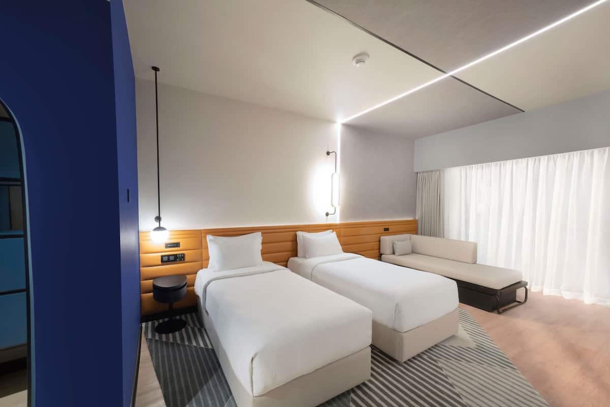 A hotel room with two beds, a sofa, a desk, and a large window with curtains.