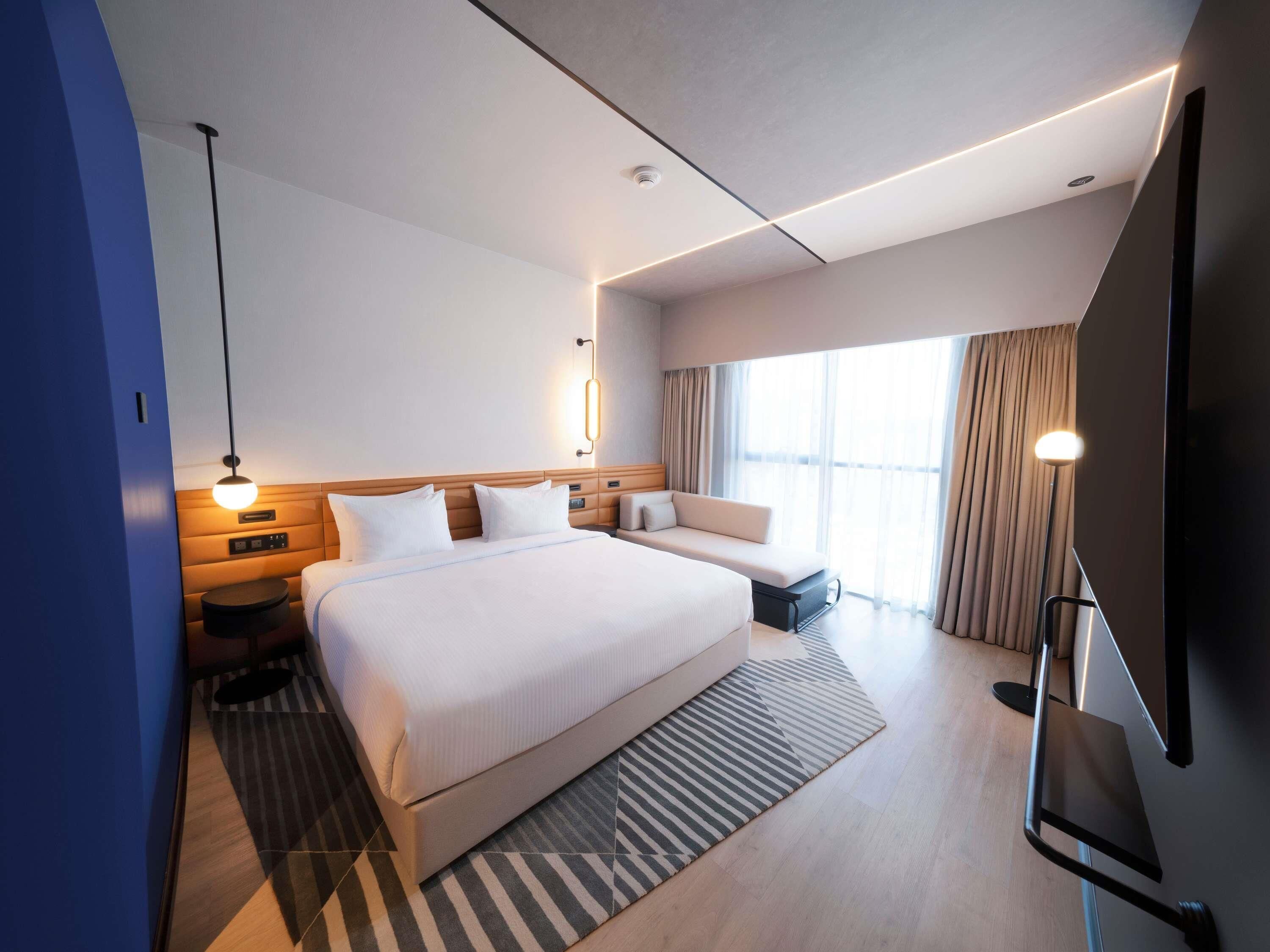 A modern hotel room with a large bed, bedside tables, a TV, and a large window with curtains.