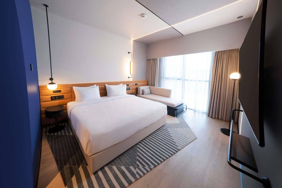 Una habitación de hotel moderna con una cama grande, mesitas de noche, un televisor y un amplio ventanal con cortinas.
