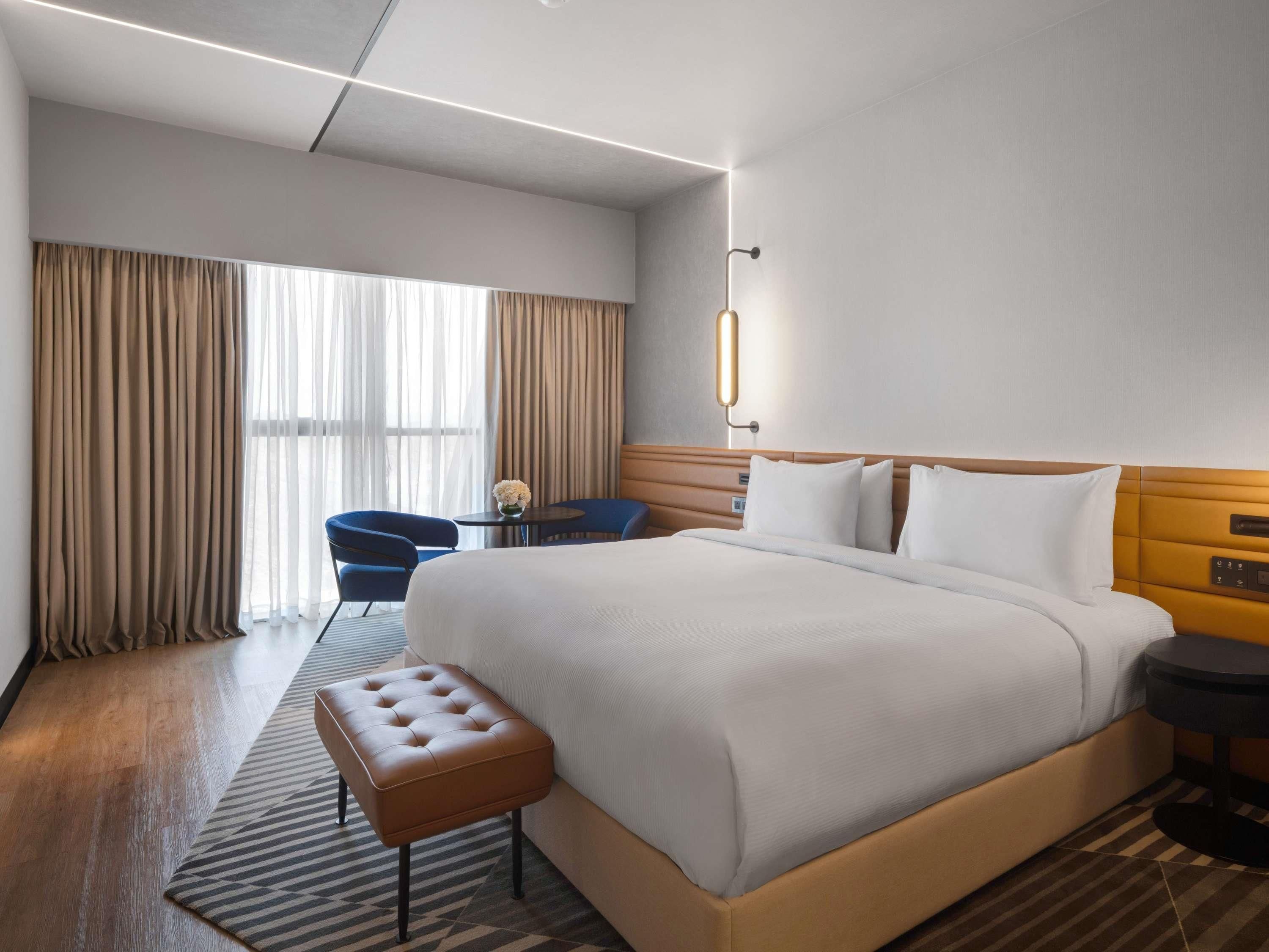 A hotel room with a large bed, a small round table, a blue chair, and a bench.