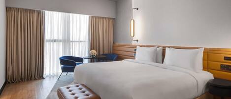 A hotel room with a large bed, a small round table, a blue chair, and a bench.