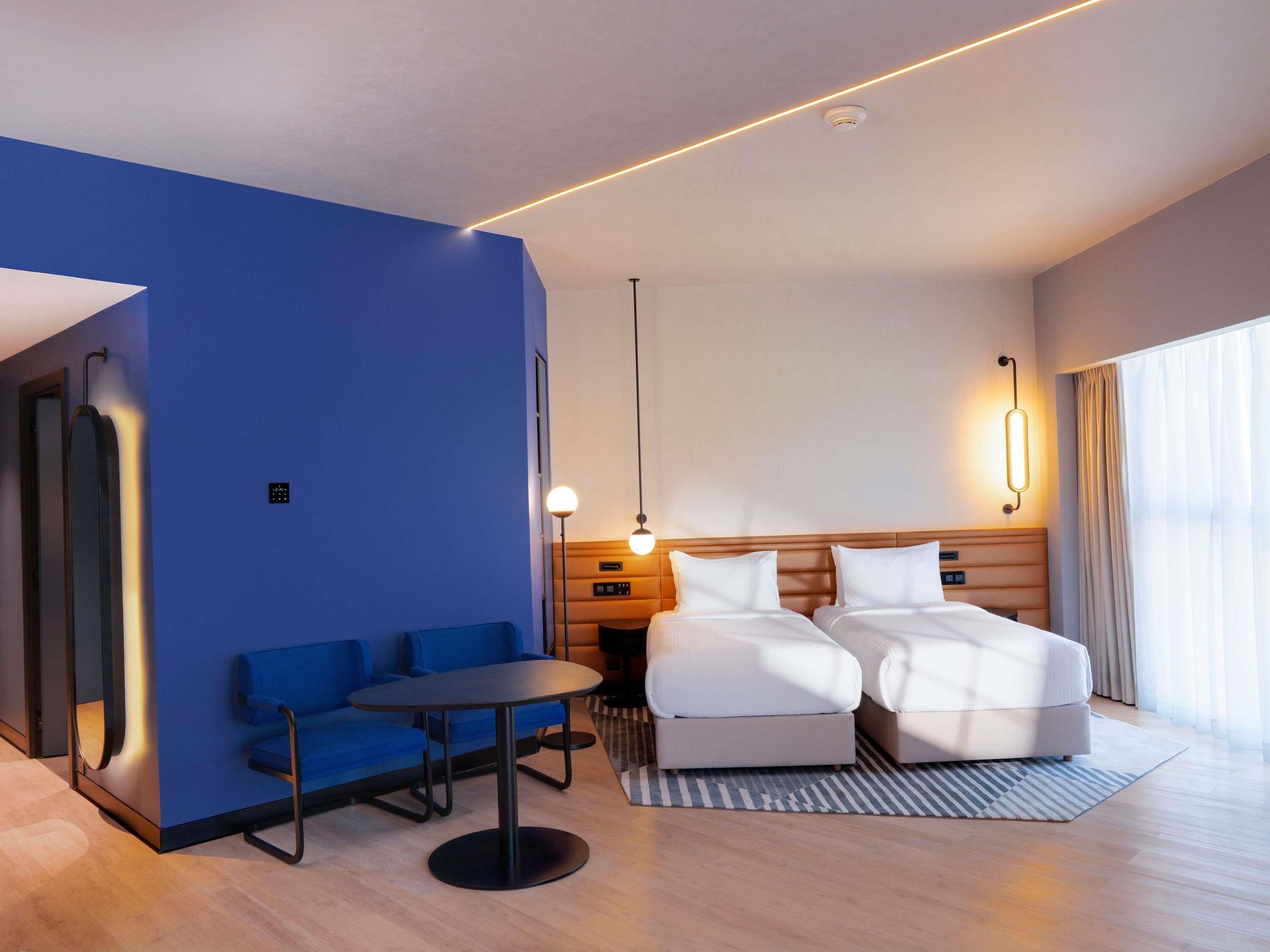 A modern hotel room with a blue accent wall, a bed with white bedding, a small round table, and a blue sofa.