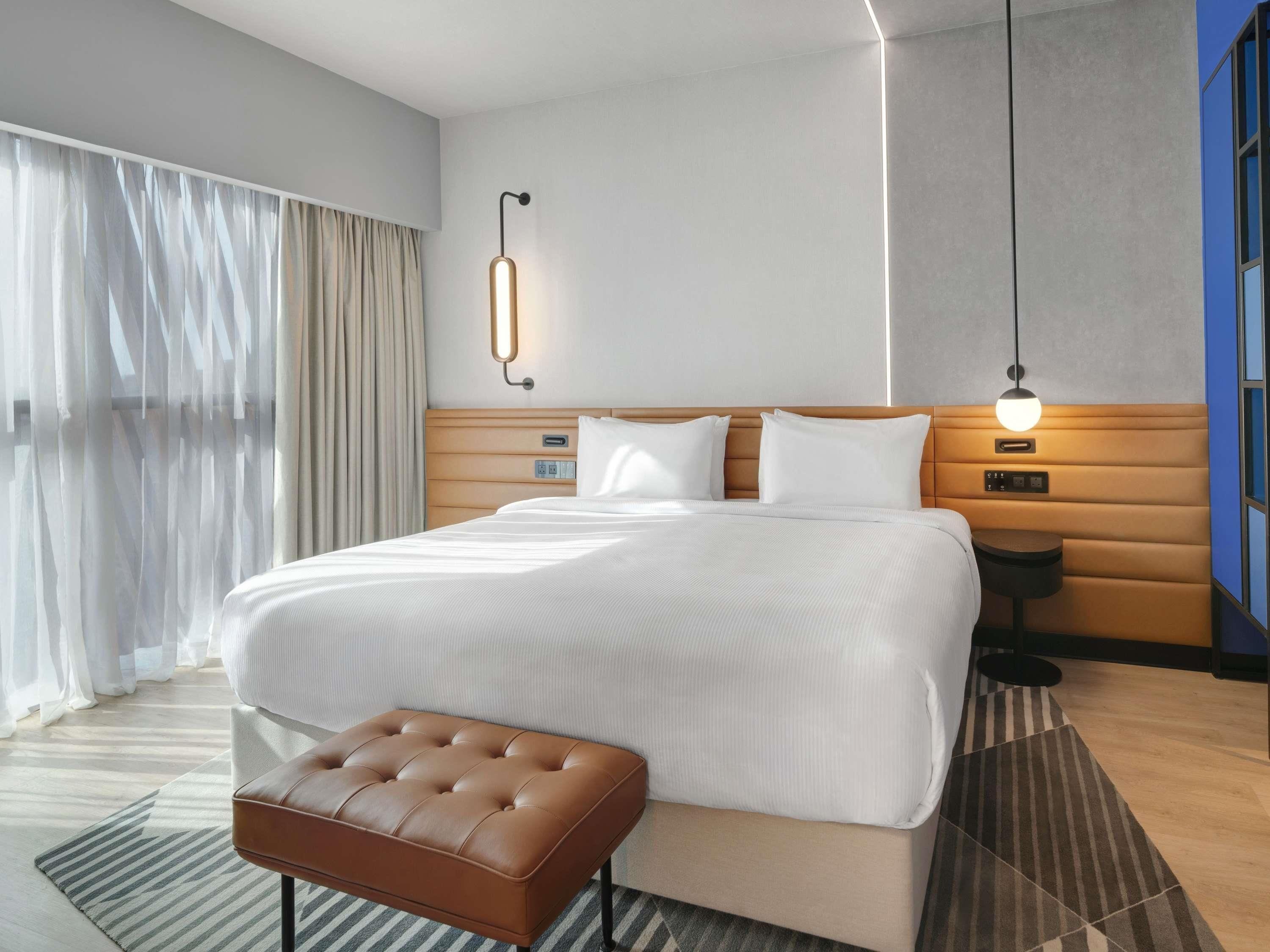 A hotel room with a large bed, bedside tables, a bench, and a view of the outside.