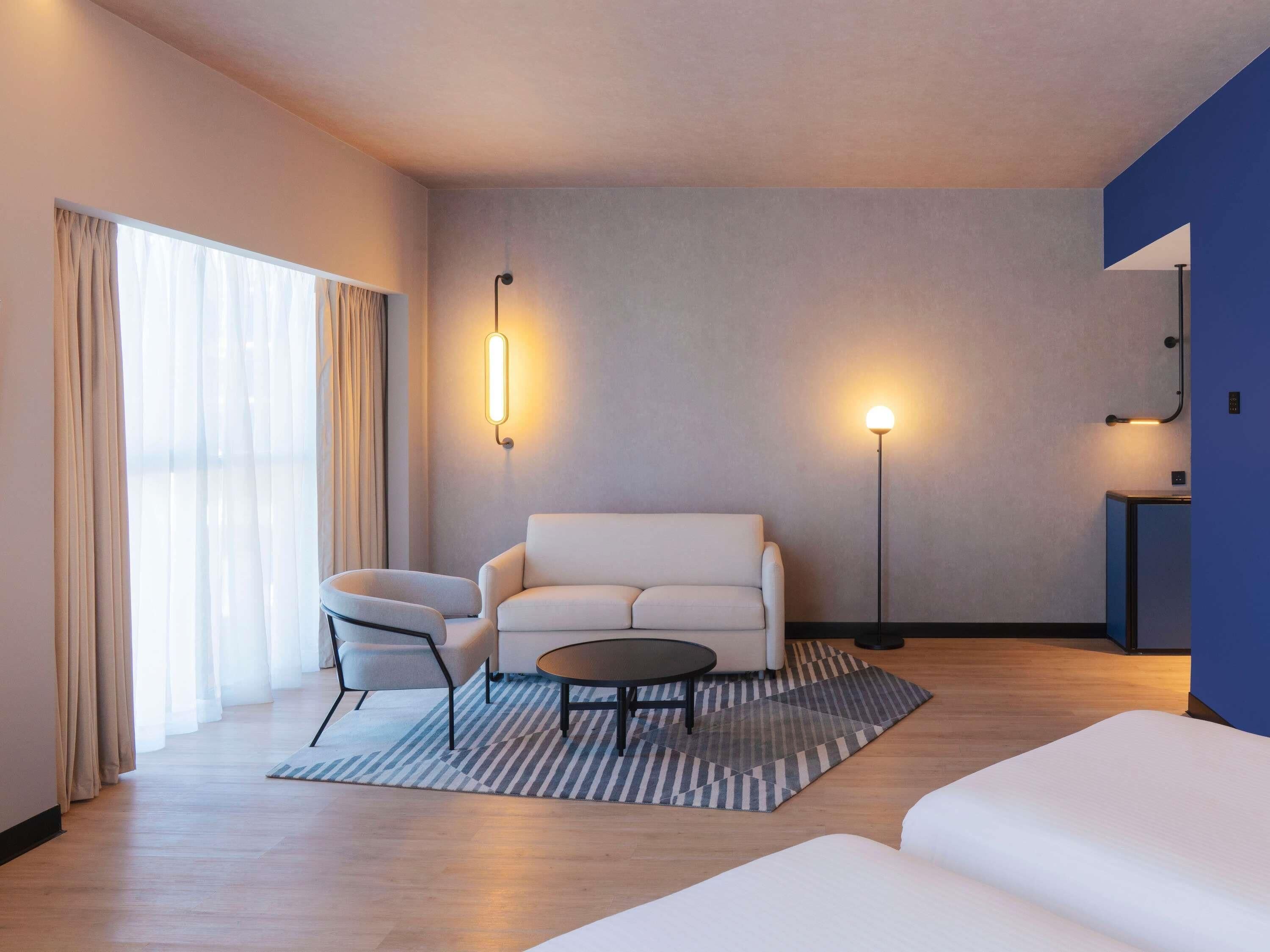 Quarto de hotel moderno com sofá, poltrona, mesa de centro redonda e escrivaninha com luminária.