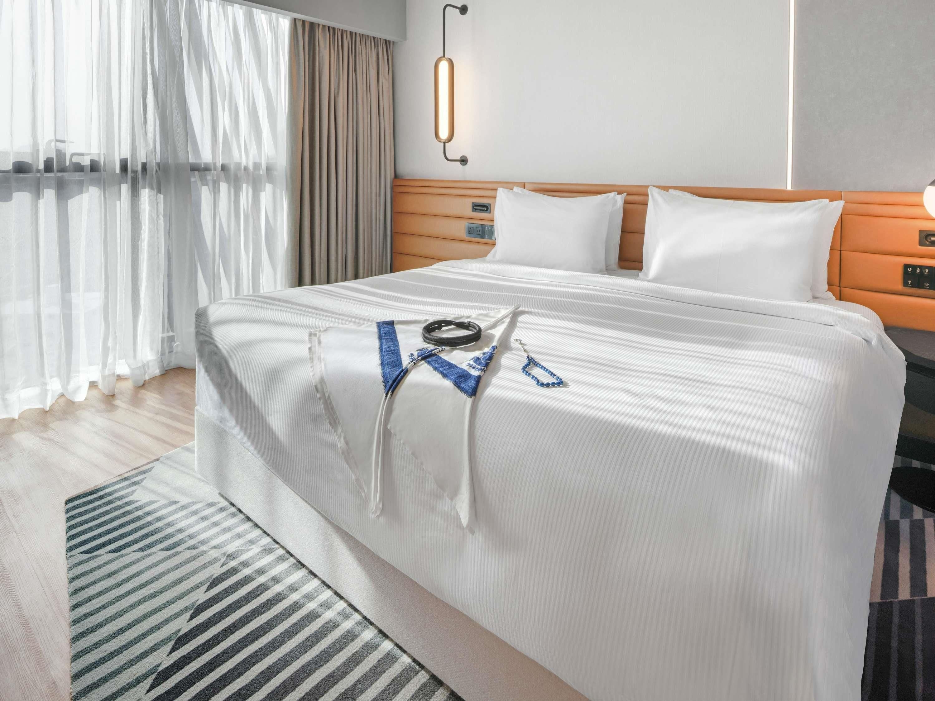Habitación de hotel con una cama bien tendida, una mesita de noche, una lámpara y un ventanal con cortinas.