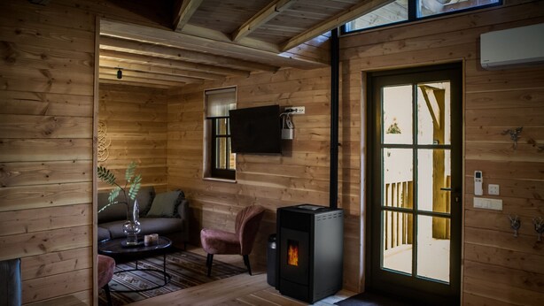 A cozy wooden cabin interior with a fireplace, a sofa, a chair, and a television mounted on the wall. - Unique overnight stay for true 'lovebirds' in our treehouse! (Uden)