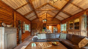 Ein Blockhaus-Interieur mit Holzbalkendecke, Kamin, Sofa, Couchtisch mit Schachspiel und einer Treppe, die zu einer Schlafempore führt.