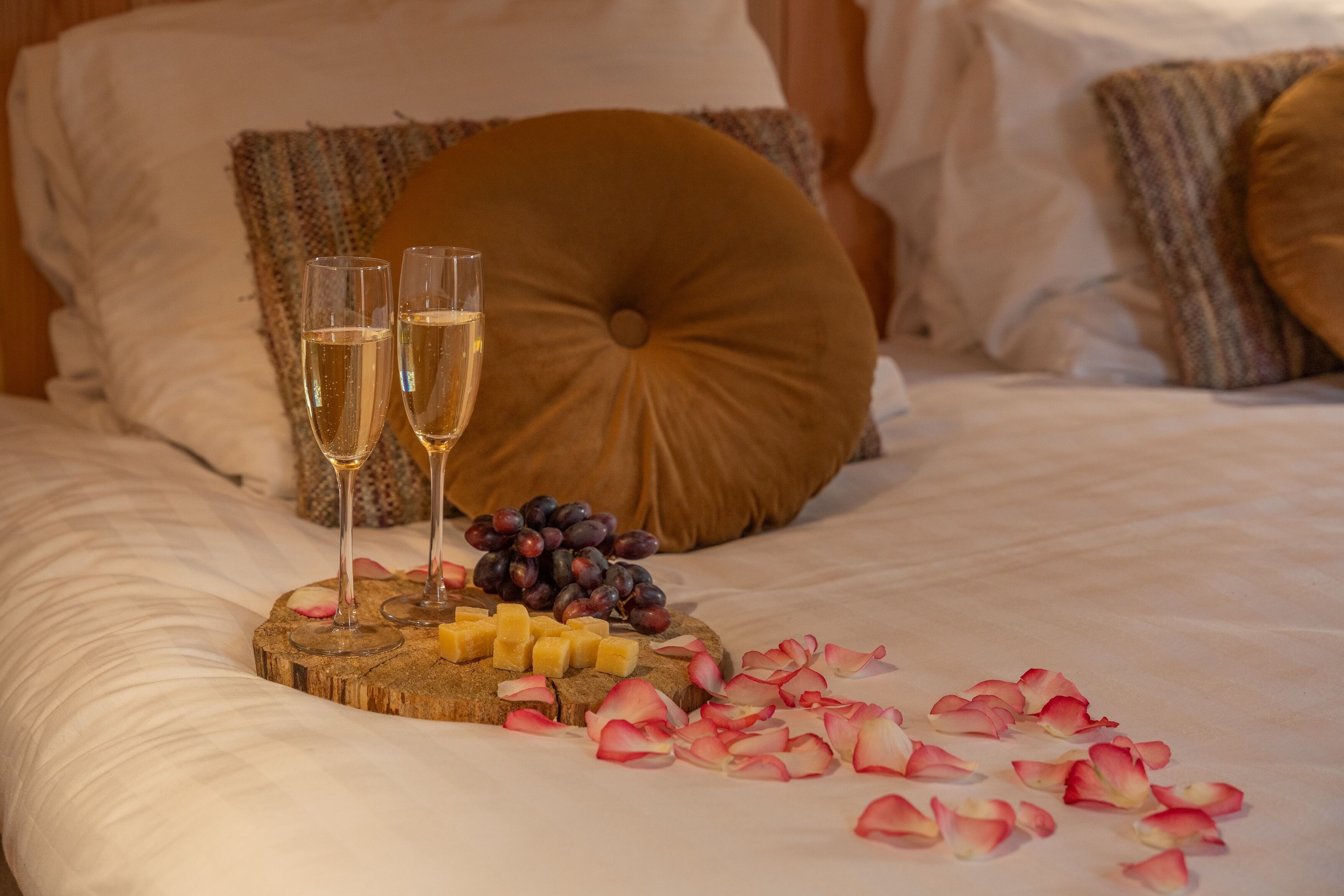 A bed with a wooden serving board holding cheese, grapes, and two glasses of champagne.