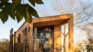 En moderne træhytte med et stort glasvindue, en træterrasse og en bænk.