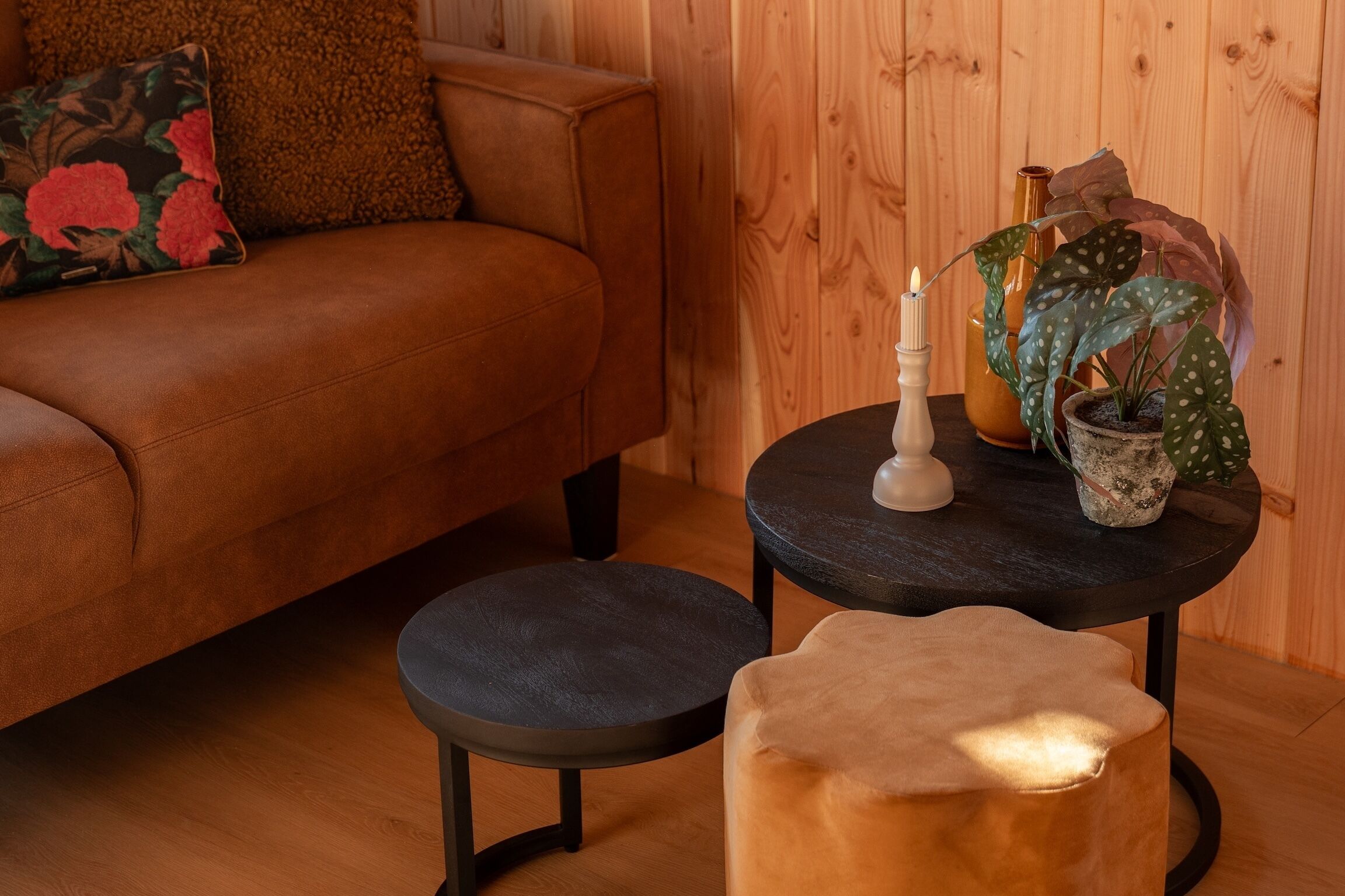 Een knus interieur met een bruine leren bank, een klein rond tafeltje met een kaars, een potplant en een houten wand.