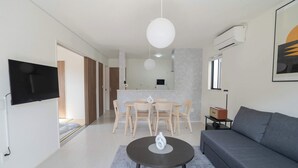 A modern living room with a sofa, a coffee table, a TV mounted on the wall, a dining area with chairs and a table, and an air conditioning unit.