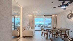 A modern living room with a sofa, dining table, and chairs. There is a large window with curtains, and a ceiling fan is visible. - Domaine Collection Sun Villa (Hersonissos)