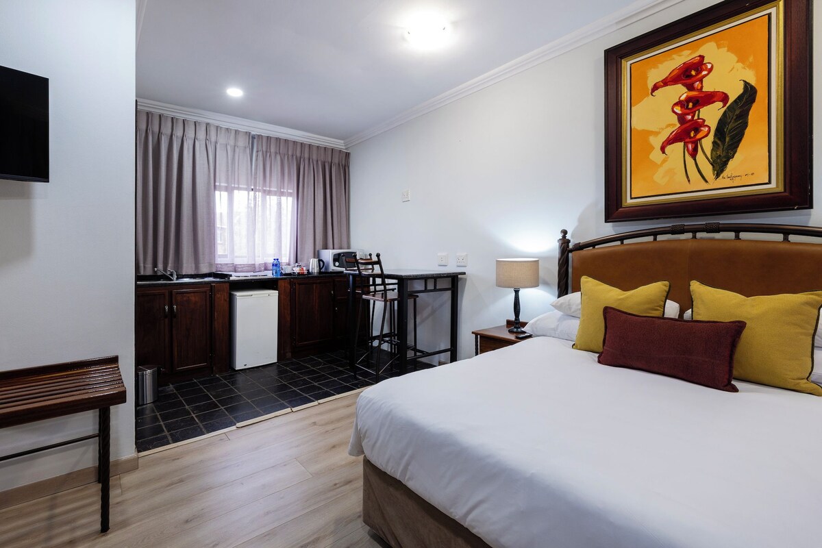 A hotel room with a bed, a desk, a chair, a lamp, a painting, and a television.