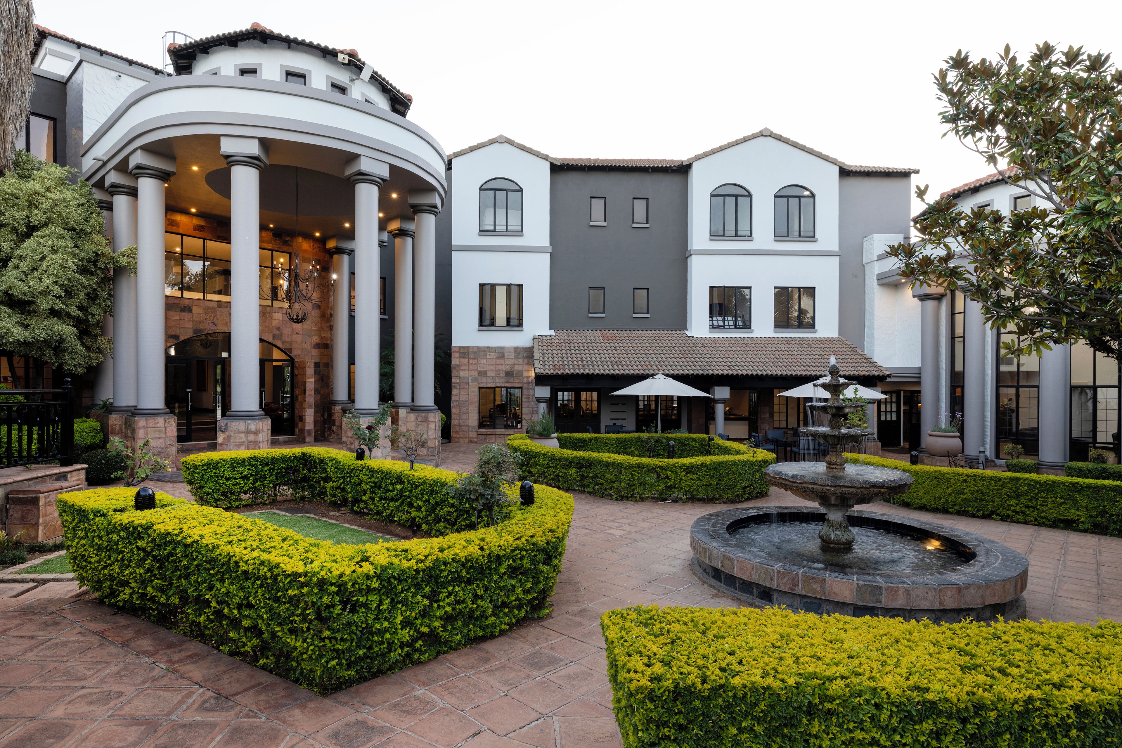 Un patio con una fuente, setos cuidados y un edificio con columnas y un porche cubierto.