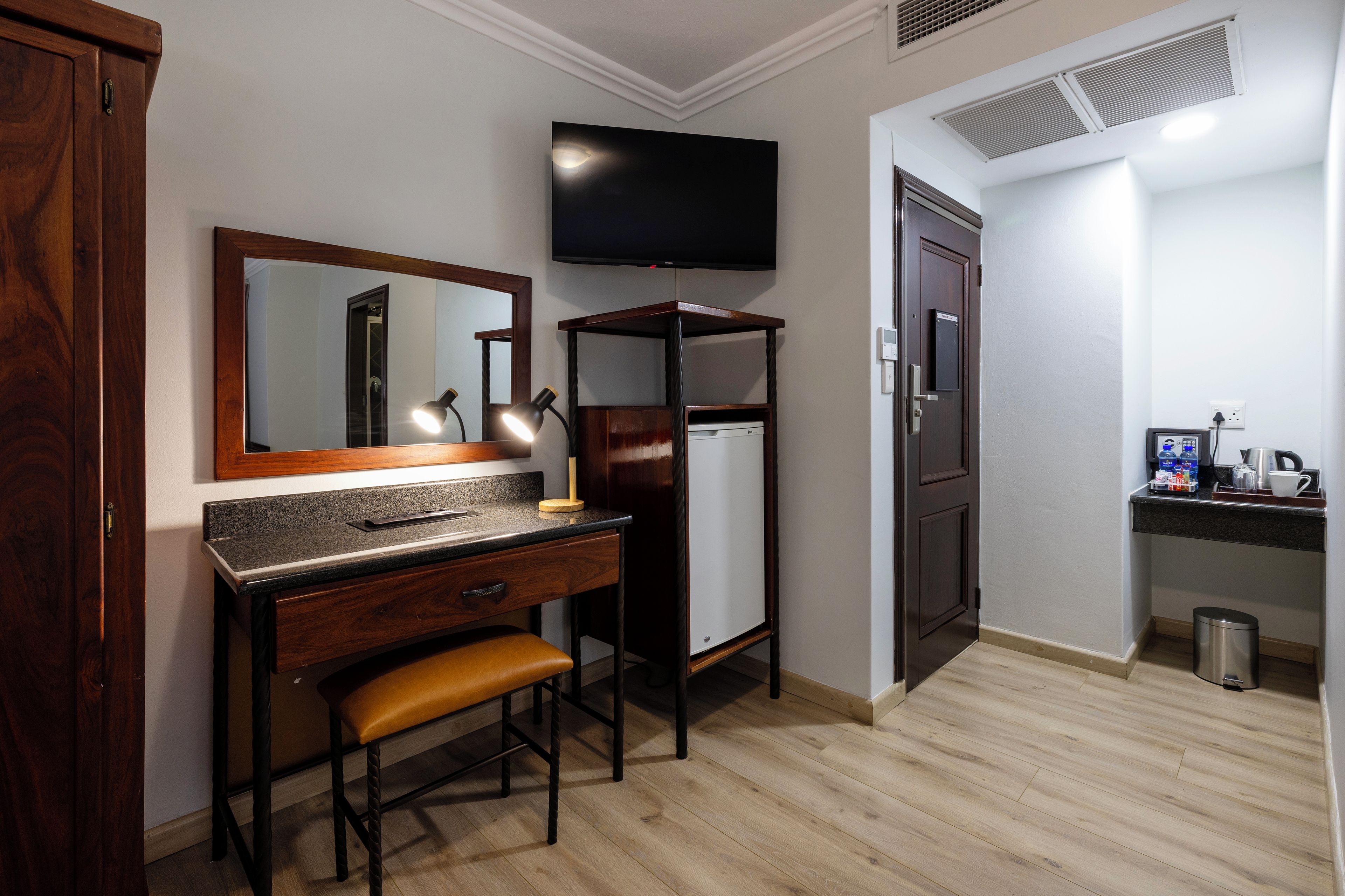 A hotel room with a wooden desk, a bench, a mirror, a television, and a kitchenette area.