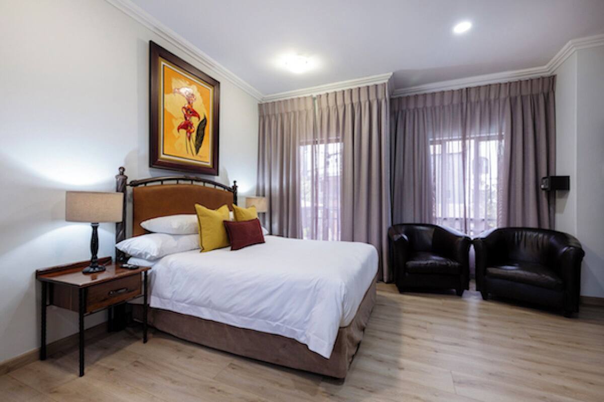 A hotel room with a bed, a nightstand, a lamp, a framed picture, two leather chairs, and a window with curtains.