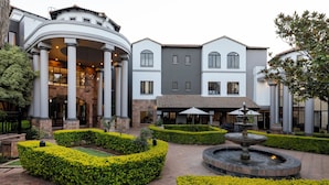 A courtyard with a fountain, manicured hedges, and a building with columns and a covered patio area.
