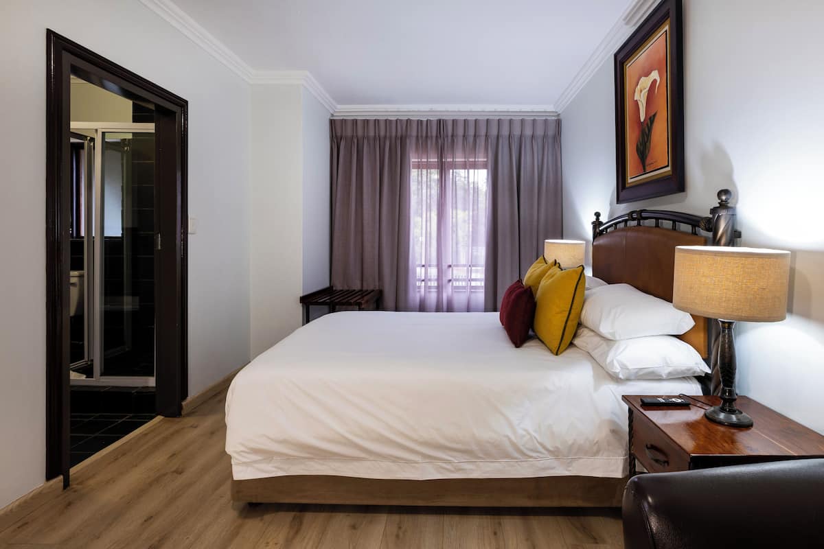 A hotel room with a bed, a nightstand, a lamp, a framed picture on the wall, and a door leading to a bathroom.